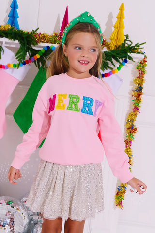 A child wearing a pink sweatshirt with 'Merry' in multicolor letters, standing in a festive setting.