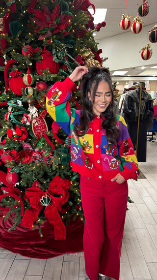 A colorful cardigan with rainbow blocks and gingerbread man motifs, button-up front, worn by a model in front of a Christmas tree.