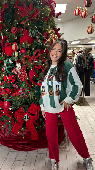 A green and ivory striped sweater with nutcracker motifs, worn by a person standing next to a decorated Christmas tree.