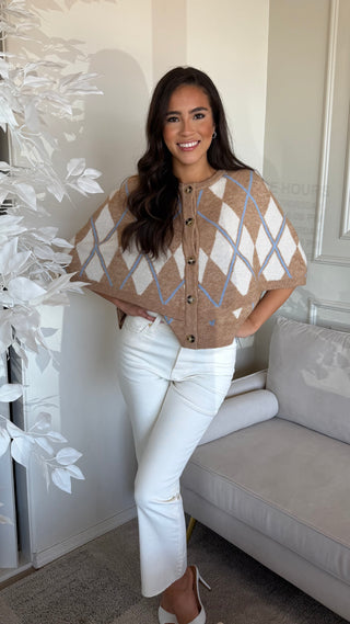 A woman wearing a beige cream argyle button-up cape sweater, paired with white jeans.
