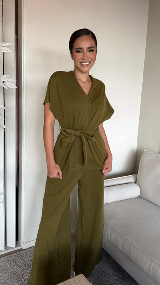 A woman wearing an olive wrap waist top and wide leg pant set, standing in a living room.