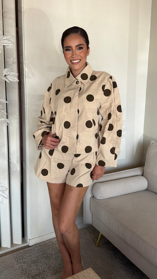 A woman wearing a beige top and shorts set with black polka dots, featuring a cowl mock neck and long sleeves.